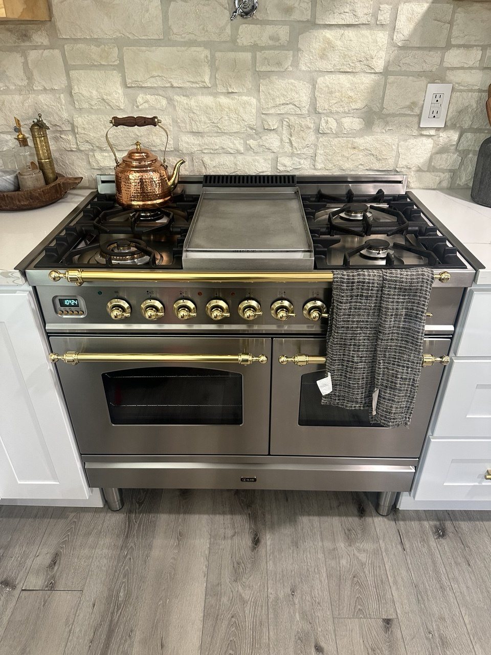 Stainless ILVE range with brass knobs and rails, a center griddle and a copper kettle on the front burner