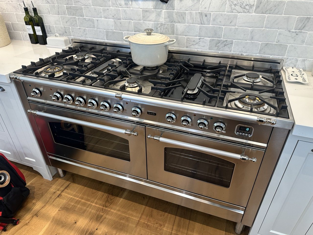 Wide stainless ILVE range with two ovens, chrome knobs and trim, and a white pot on the cooktop